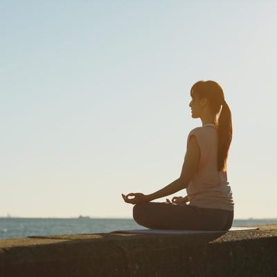 A woman sits cross legged by the ocean practicing mindfulness techniques from her online mindfulness therapy